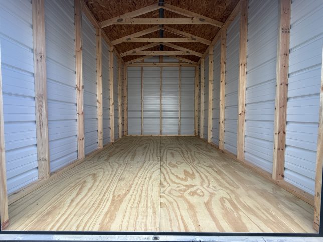 Interior view of an 8×12 metal shed showing exposed wood wall studs, roof trusses, metal siding, and treated plywood floor.