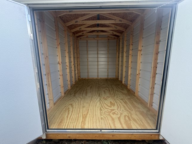 Interior of an 8×12 metal utility shed showing treated plywood flooring, exposed wood framing, and roof trusses through open double doors.