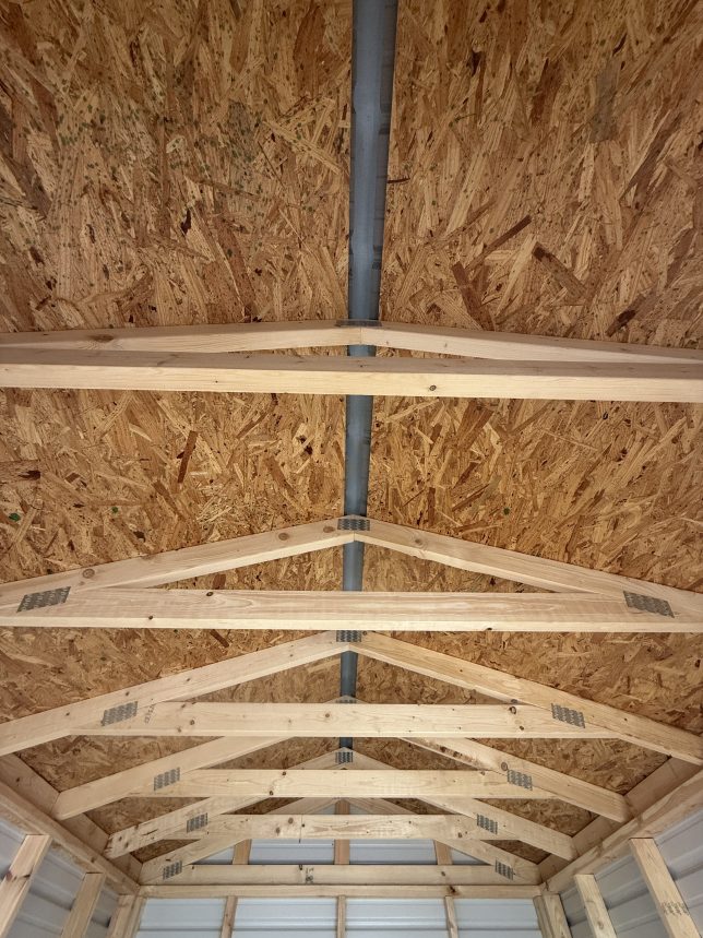 Close-up of wooden roof trusses with metal connector plates and the ridge ventilation channel inside an 8×12 utility shed.