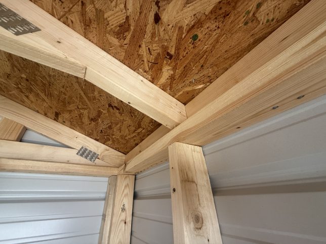 Close-up of a wood roof truss bearing on the wall framing and strongback bandboard inside a metal utility shed.